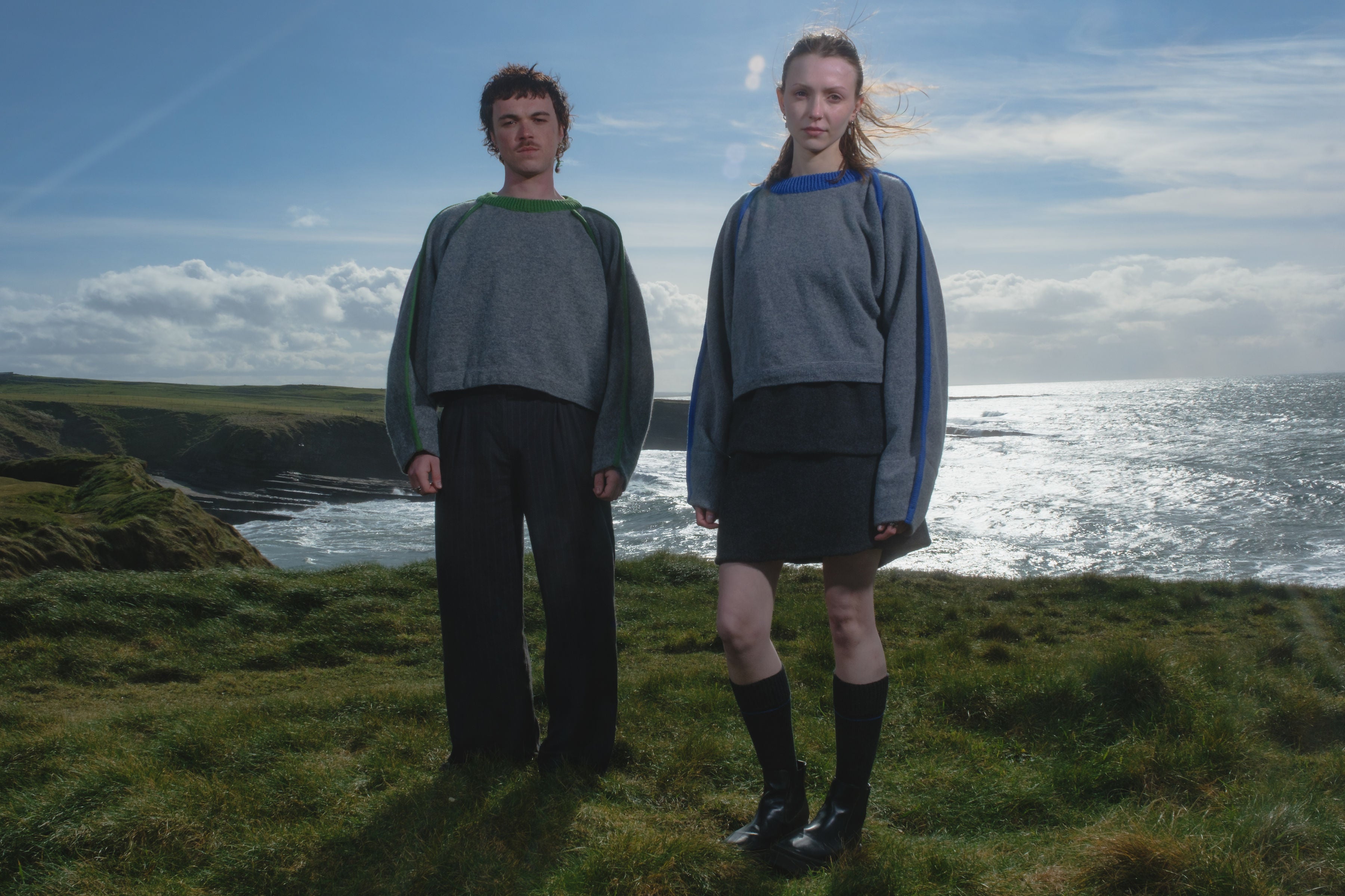 Two people wearing lambswool jumpers in grey with contrast piping, standing on a grassy cliff overlooking the ocean with a blue sky.