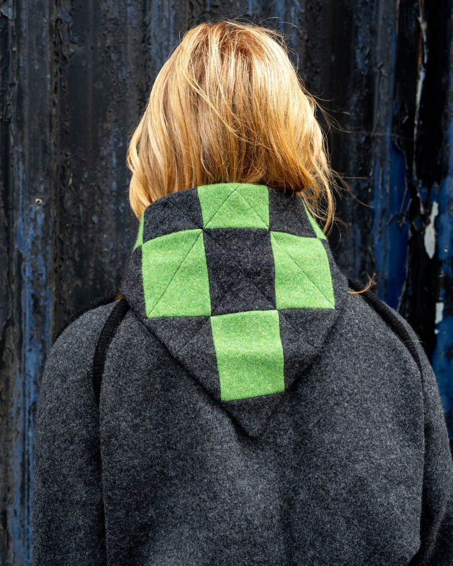Person wearing a hoodie with a green and black checkered pattern on the back, standing against a dark wall.