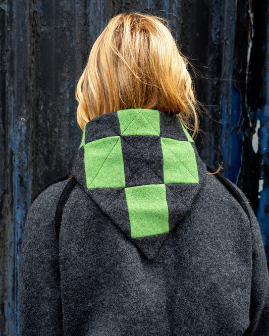 Person wearing a hoodie with a green and black checkered pattern on the back, standing against a dark wall.